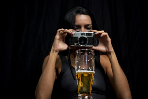 工作室肖像的年轻女子，在黑色的t恤，她的脸上有一个相机和一杯啤酒，在黑色的工作室背景。图片下载