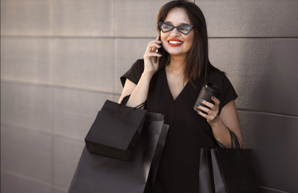 美丽自信的大码女人穿着黑色连衣裙，戴着现代眼镜，喝着咖啡，在黑色星期五大减价后拿着黑色购物袋打电话。消费主义的概念图片下载