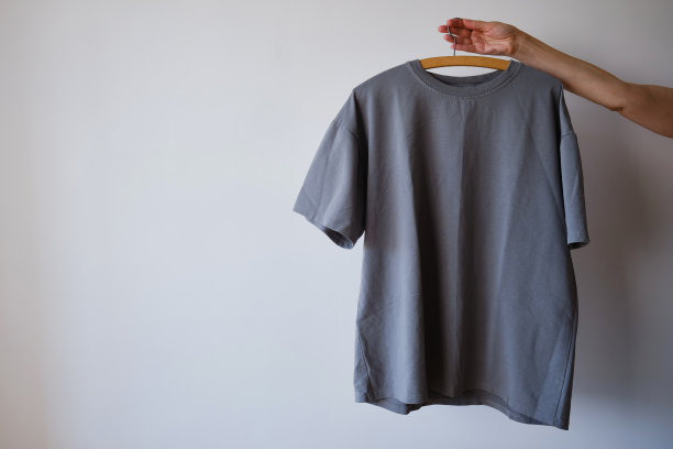 灰色t恤挂在衣架上，手里拿着浅色背景。复制空间。衣架上的衣服图片下载