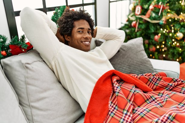 年轻的非裔美国人放松，手放在头上坐在沙发旁的圣诞树在家里。图片下载