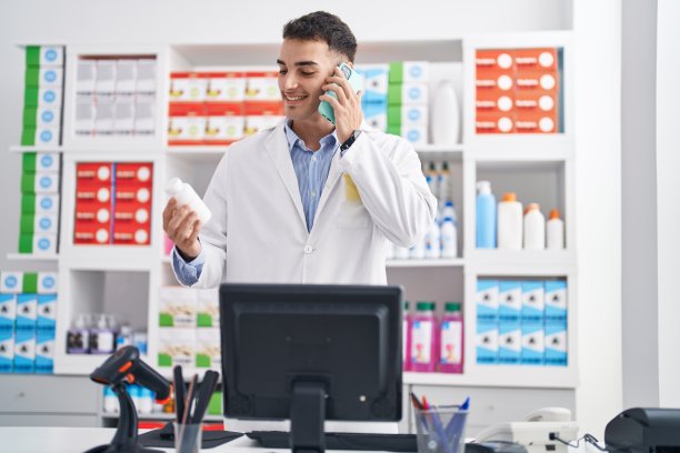 年轻的西班牙男子药剂师拿着药片在药店用智能手机说话图片下载