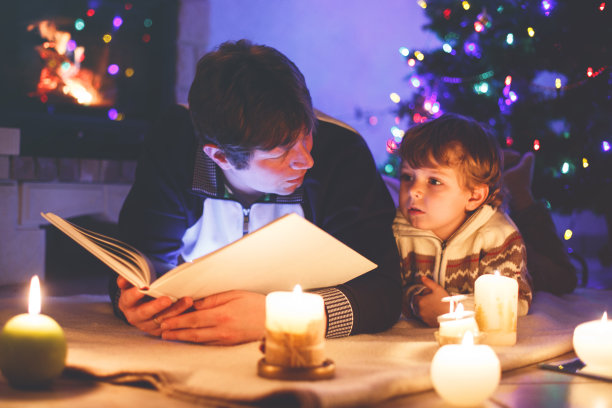 父亲和可爱的小男孩在烟囱、蜡烛和壁炉旁看书。图片下载