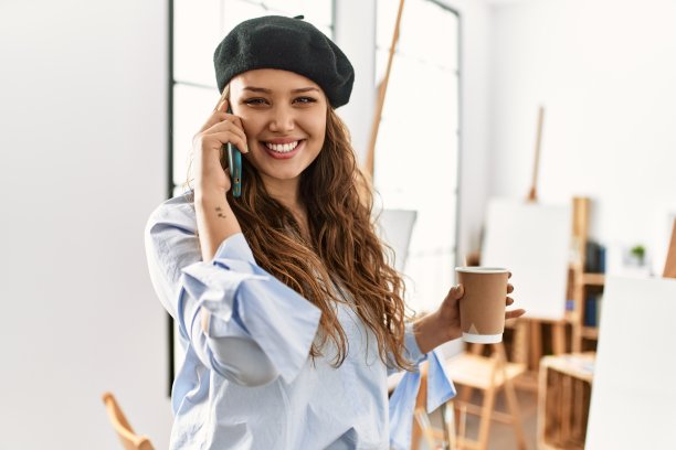 年轻美丽的西班牙女艺术家在艺术工作室喝咖啡，用智能手机说话图片下载