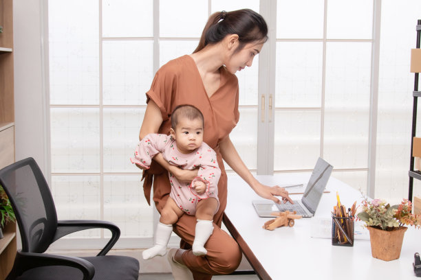 美丽的亚洲妈妈抱着蹒跚学步的孩子，试图在家里用笔记本电脑工作，妈妈和孩子一起在电脑前工作。新生的粉红色棉软纺织樱桃图案。育儿和工作母亲的概念。图片下载