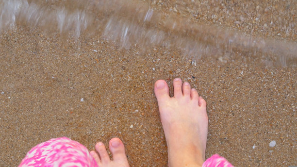 俯视图女性的腿，被海浪冲刷。一个穿着衣服的女人站在岸上，没有穿鞋，她的脚踩在潮湿的沙子上，沙子在水里。海滨度假的概念。热带旅行癖图片下载