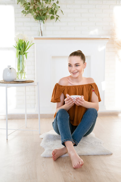 年轻女子坐在房间与装饰壁炉在地板上与马克杯在她的手图片下载