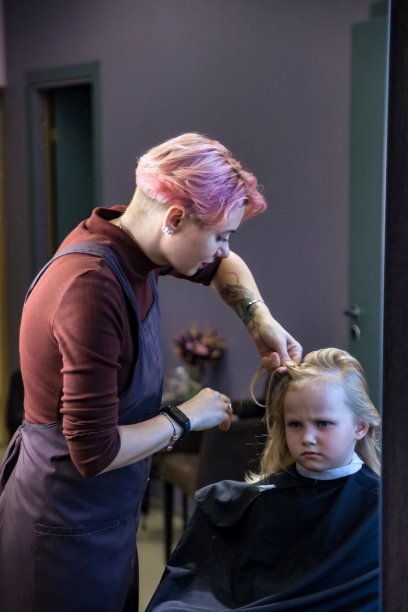 美发师妇女在理发店为可爱的金发小女孩做时髦的发型。理发师在理发店、发廊为宝宝做有风格的发型。发型，美的概念。本空间图片下载