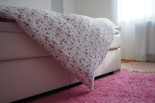 舒适紧凑的卧室，白色家具，粉色地毯和薄纱窗帘。双人床上盖着毛茸茸的羽毛毯子。浪漫的早晨。窗外的光线。毯子垂到地板上。图片下载