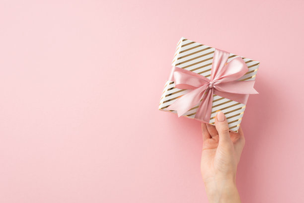 圣诞夜庆祝概念。第一人称俯视图照片的妇女手握时尚的礼物盒与粉色丝带蝴蝶结上孤立的浅粉色背景与copyspace图片下载