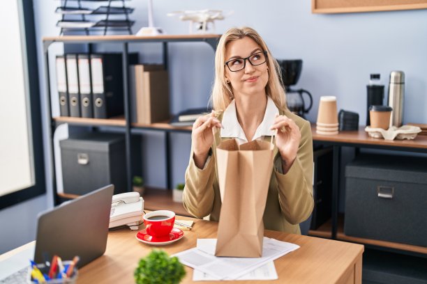 年轻的白人妇女在办公室里拿着纸袋微笑着看着一边，盯着别处思考。图片下载