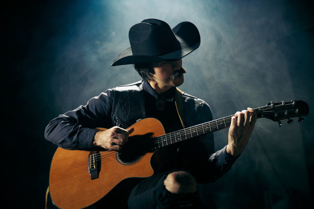 肖像男子留着小胡子，穿着乡村风格的衣服，弹奏吉他，在深蓝色背景与烟雾隔离图片下载