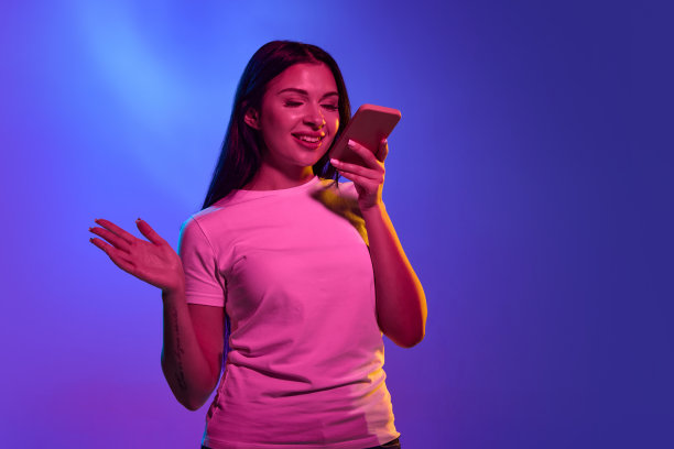 用着手机，惊呆了。在霓虹灯下，白人妇女的肖像被孤立在渐变的工作室背景上。漂亮的女模特。人类情感的概念，面部表情，销售，广告。图片下载
