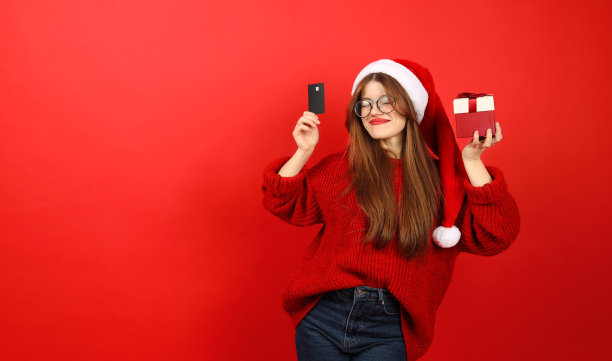 圣诞节的折扣。一个戴着圣诞帽、穿着节日毛衣的快乐女孩，手里拿着购物用的信用卡和红色背景的礼物。图片下载