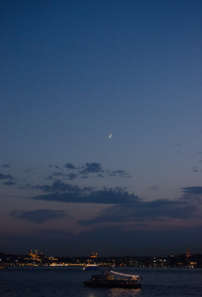 垂直拍摄一艘船在一个城市的水体下，夜空下有半月图片下载