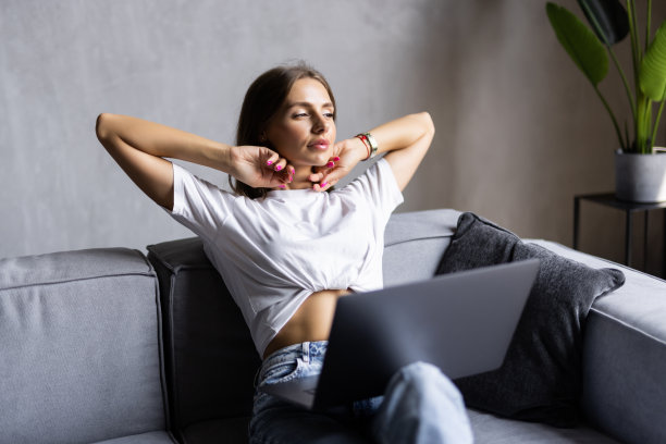 一名穿着白衬衫的年轻女子拿着笔记本电脑坐在沙发上，在工作中坐了很长时间后，她把手臂举过头顶伸展开来。图片下载