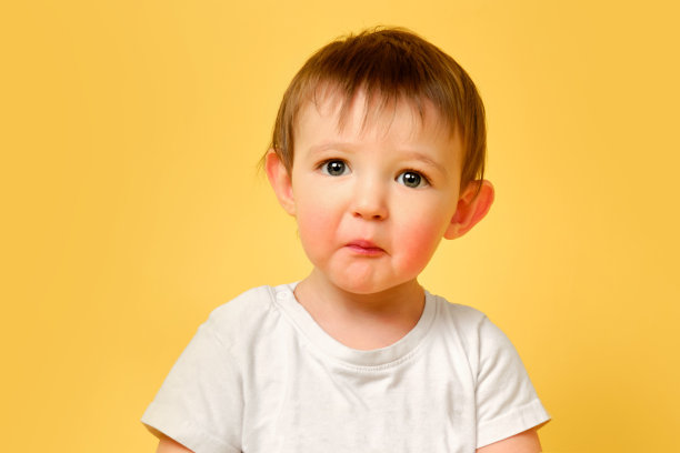 一个心烦意乱的蹒跚学步的婴儿与一个惊讶的悲伤的脸在工作室黄色的背景。头发蓬乱，穿着白t恤的冒犯儿童。一岁零四个月的孩子图片下载