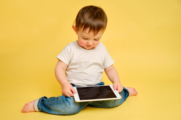 蹒跚学步的婴儿在录音棚黄色的背景上用数码平板电脑。穿着白t恤和蓝色牛仔裤玩平板电脑的快乐孩子图片下载