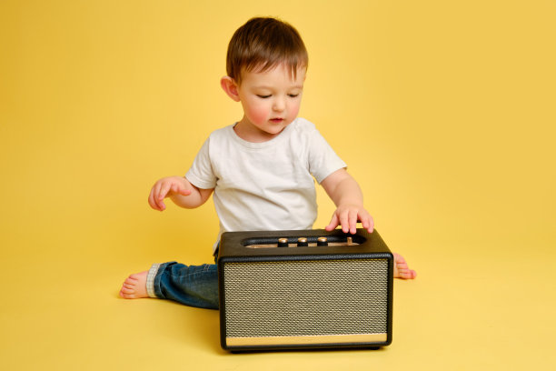蹒跚学步的婴儿在录音棚的黄色背景上玩无线音乐扬声器。穿着白t恤和蓝色牛仔裤的快乐孩子在听扬声器里的音乐。一岁零四个月的孩子图片下载
