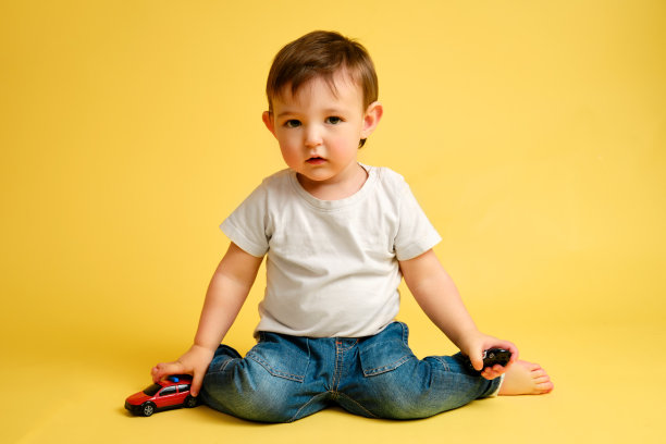 一个蹒跚学步的婴儿在工作室的黄色背景下玩玩具汽车。穿着白t恤和蓝色牛仔裤的快乐孩子在玩玩具汽车。一岁四个月大的孩子图片下载