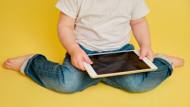 蹒跚学步的婴儿在录音棚黄色的背景上用数码平板电脑。穿着白t恤和蓝色牛仔裤玩平板电脑的快乐孩子。一岁零四个月的孩子图片下载