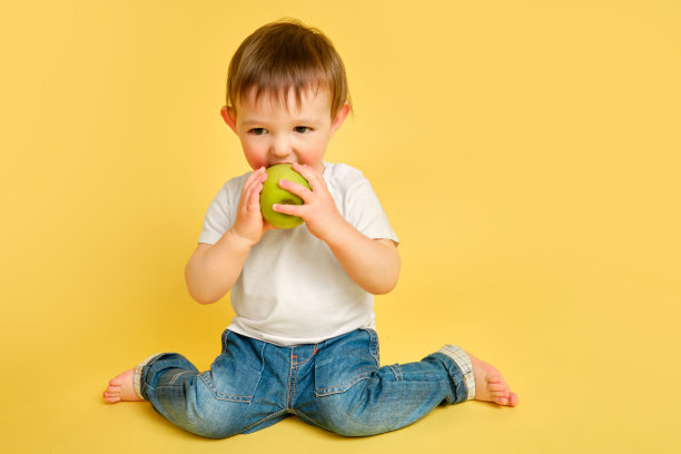 快乐的蹒跚学步的婴儿在工作室的黄色背景上吃着一个绿苹果。穿着白色t恤和蓝色牛仔裤，带着一个大绿苹果的孩子，模仿空间。一岁零四个月的孩子图片下载