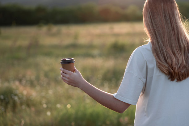 女人的手拿着纸工艺咖啡杯在大自然美丽的夕阳下。图片下载