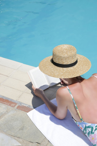 美丽的年轻女子的肖像享受炎热的一天阅读一本书旁边的游泳池。放松的暑假概念。图片下载