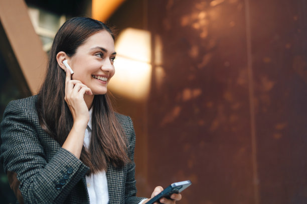 迷人的商务女性戴着耳机微笑着看向别处站在办公楼前面。远程工作、商务、自由职业、网上购物、在线学习、城市概念。副本的空间。图片下载
