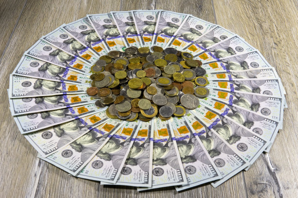 一百美元的美钞摆成一个圈，中间是一堆来自世界各地的硬币，放在灰色的木头上图片下载