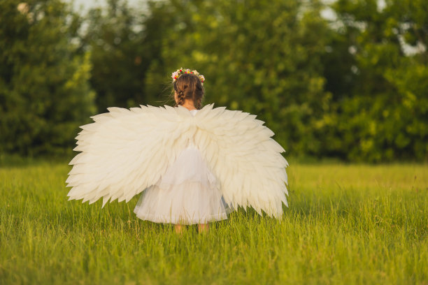 绿色草地上长着白色翅膀的天使。金发女孩在夏天的背景下穿着衣服图片下载