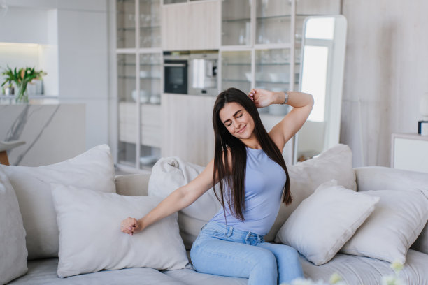 可爱苗条运动的年轻女子松散的深色头发伸展眼睛闭着，坐在客厅舒适的沙发上。快乐的家庭主妇在家享受星期天的早晨。国内舒适休闲。图片下载
