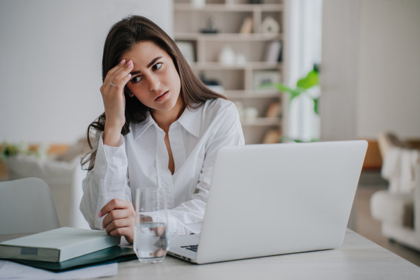 筋疲力尽的年轻黑发女孩坐在桌子旁，拿着笔记本电脑、日记、纸和一杯水，靠在她的手看向另一边，感到她的工作焦虑。疲惫的学生对着模糊的内心。图片下载