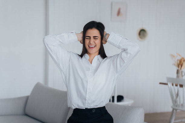 沮丧的女人穿着白衬衫黑裤子坐在家里的沙发上，挤压着头，因为可怕的消息感到难过，尖叫着闭上眼睛。女孩感到头痛，抬起头，经历强烈的疼痛。图片下载