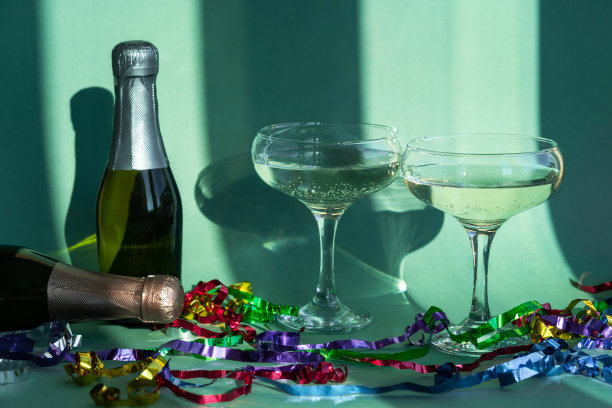 两杯香槟和几瓶香槟在阳光的阴影下，圣诞装饰在绿色的背景。庆祝新年和圣诞节。图片下载