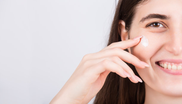 年轻女子面部特写，使用软化霜。美丽的女孩在她的脸颊上涂抹润肤霜，隔离在白色背景与复制空间。美容护肤、化妆品、spa程序概念图片下载