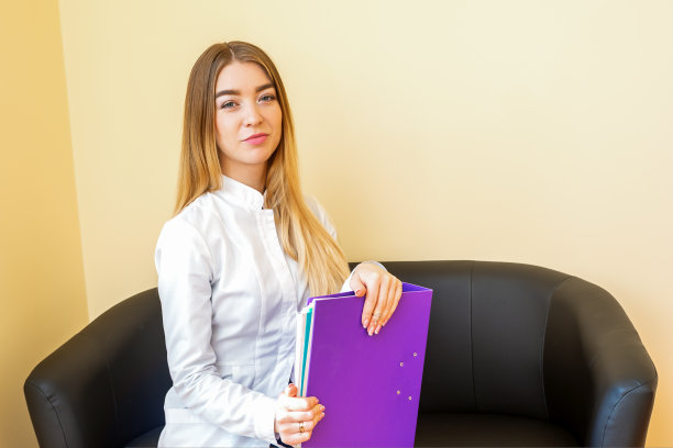 一个年轻的白人医生的肖像，拿着文件夹，金发长发，穿着白色外套，坐在黑色沙发上看着相机。图片下载