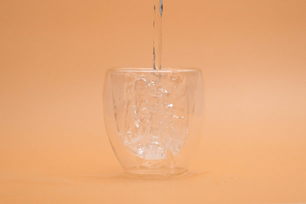 在复古背景的玻璃中倒入温水。把水倒进双层玻璃的镜头，隔离在米色背景上。图片下载