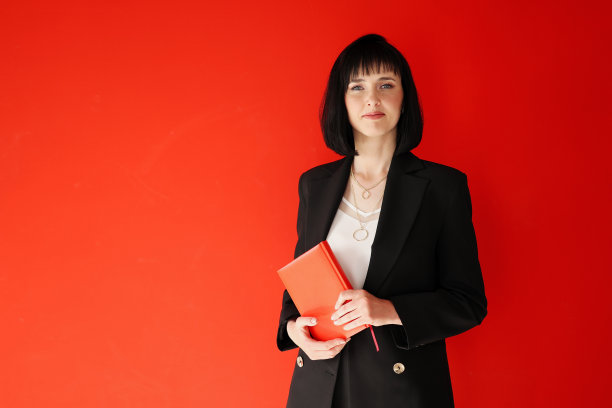 美丽的商业妇女拿着纸笔记本孤立的红色背景。成功的黑发女性，黑色套装，短发看着镜头。创业，事业成功，教育理念。图片下载