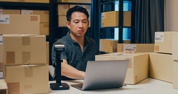 亚洲商人使用条形码机扫描纸盒中的客户数据，在笔记本电脑上输入在线注册信息，订单明细，在仓库发送快递。图片下载