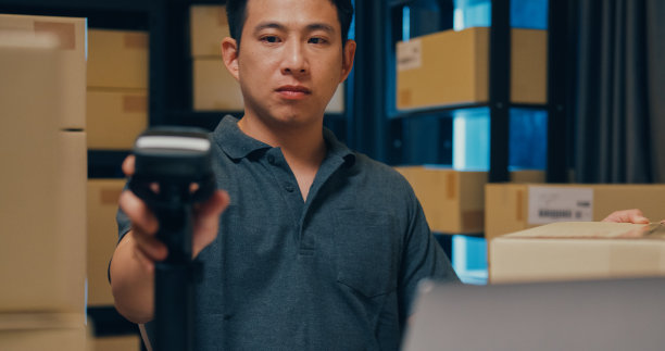 近距离的亚洲商人使用条形码机器扫描纸盒中的客户数据，并在笔记本电脑上键入注册在线信息订单明细，以便在仓库快递。图片下载