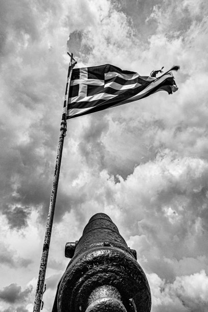 在多云的天空下，希腊国旗在古老的柱子附近的低角度灰度图片下载