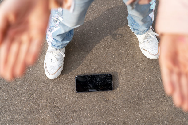 手机损坏，触摸屏破裂。脚和手的高架视图，破碎的屏幕智能手机掉在沥青概念的地板上。图片下载