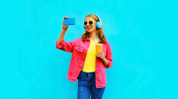 肖像快乐微笑的妇女与智能手机在无线耳机听音乐穿着粉红色夹克在鲜艳的蓝色背景自拍照图片下载