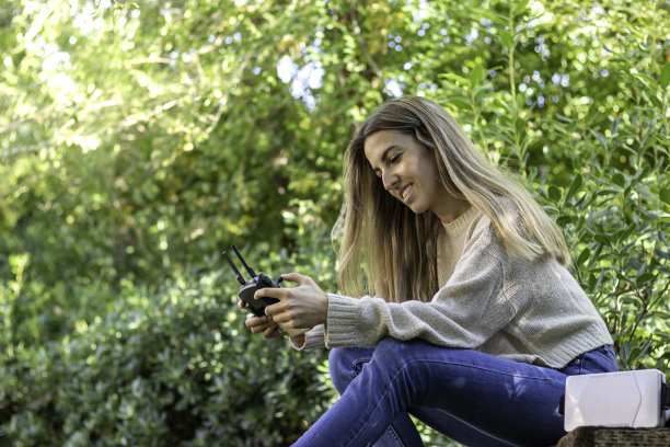 低角度的视角年轻的金发女子微笑和快乐，而飞行无人机与遥控器在手中，而在自然图片下载