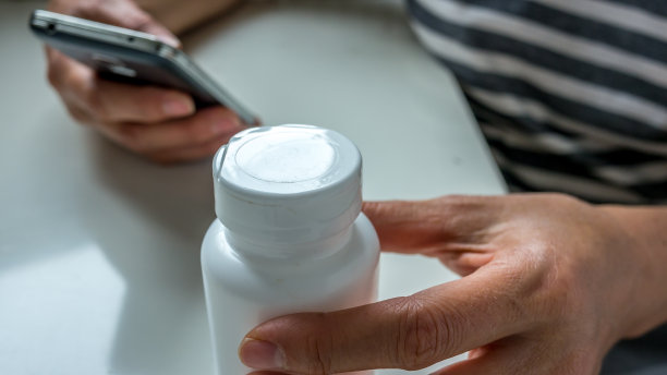 一手拿着智能手机，阅读药品说明书。另一只手拿着一个药瓶。关闭了。图片下载