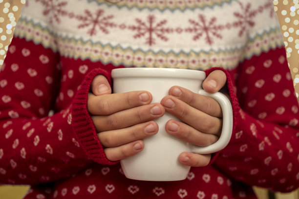 女孩穿着圣诞图案的羊毛衫，拿着一杯咖啡、巧克力或茶。圣诞彩灯在背景中散焦。图片下载