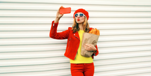 肖像时尚的年轻女子自拍与智能手机拿着杂货店购物纸袋长白色面包头戴红色贝雷帽，心形太阳镜在白色背景图片下载