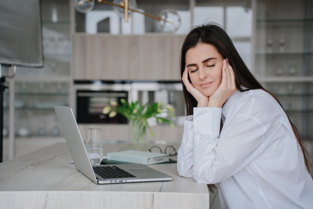 昏昏欲睡的褐发年轻女子闭着眼睛坐在桌子前，用手提电脑靠在手上，试图放松。沉思的白人女学生在网上远程课程后休息。教育和商业。图片下载