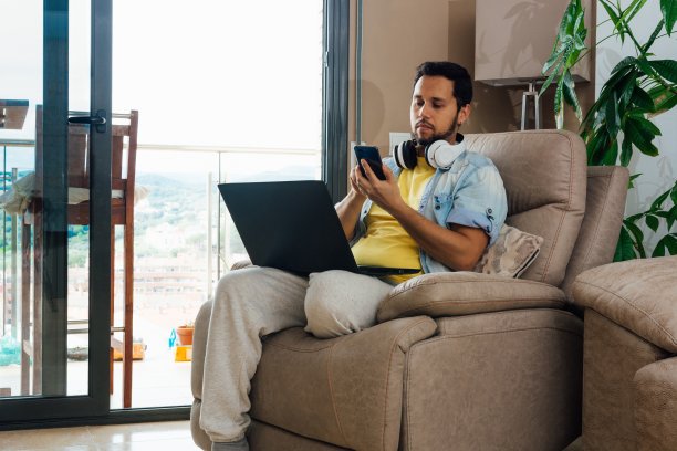 戴着耳机的年轻人在家里看手机，膝上放着笔记本电脑图片下载
