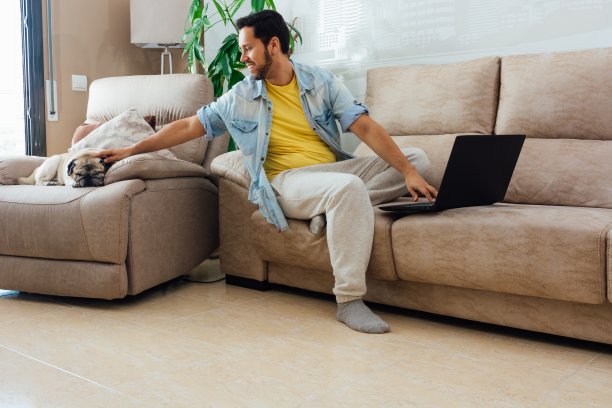 穿着休闲装的年轻人抚摸着他的狗，用着笔记本电脑，在家办公的概念图片下载